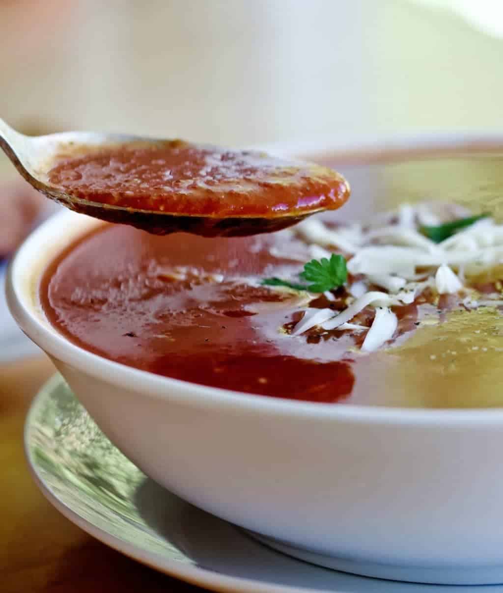 Roasted Pepper Tomato Soup shown in a white bowlGarnished with pepper jack cheese and parsley. A spoonful is lifted to see the texture.