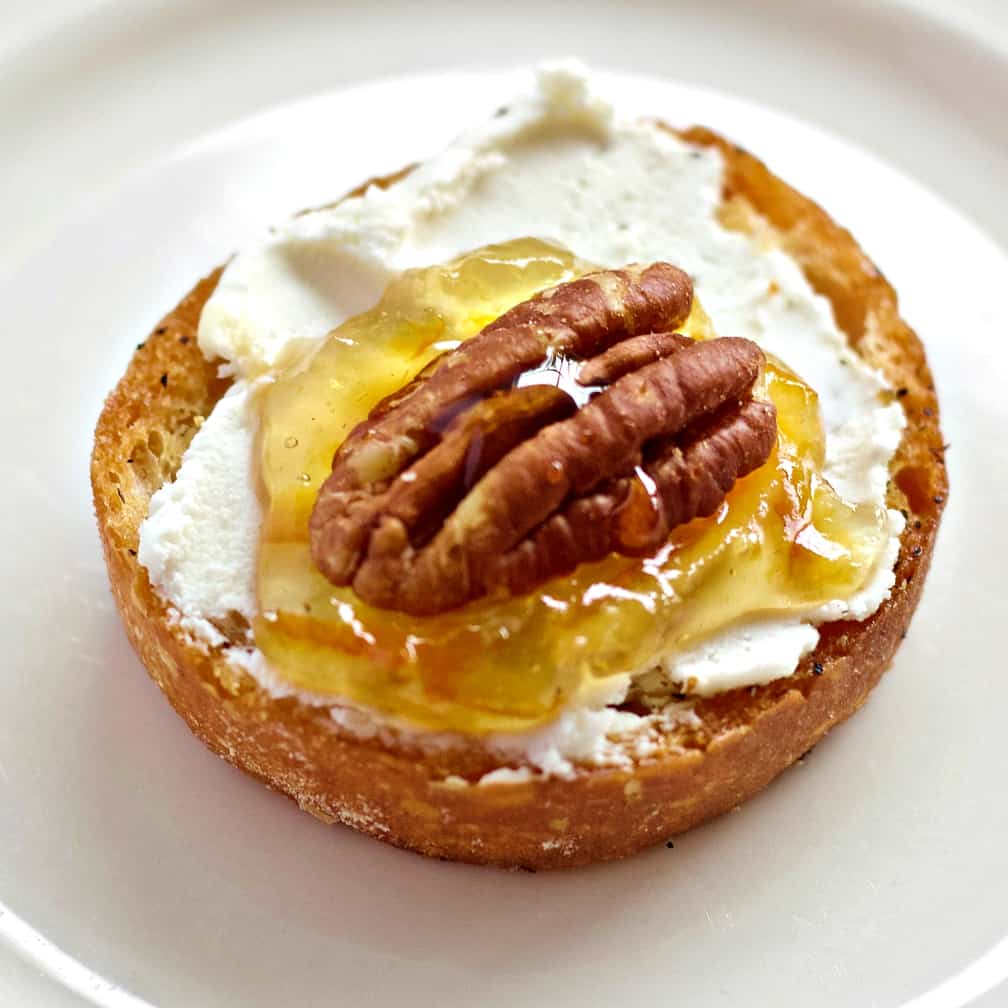 Goat Cheese Crostini Appetizer with Marmalade and Pecan 2 a single goat cheese crostini topped with orange marmalade and a whole pecan. Honey is drizzled on top. Sitting on a white plate.