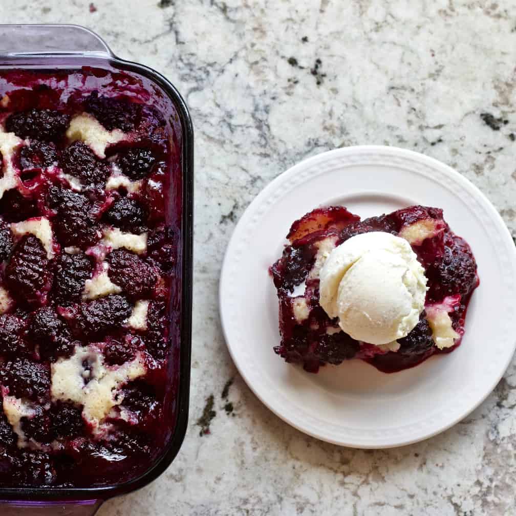 Blackberry cobbler single serving with vanilla bean ice cream sitting by a pan of this recipe.