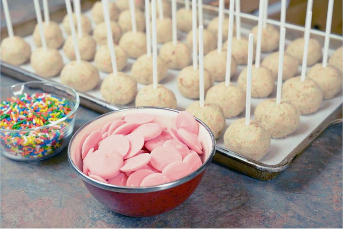 cake pops ready to dip and pink candy melts with a bowl of sprinkles.