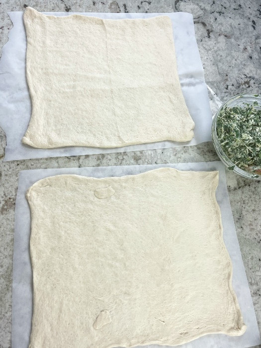 two pizza crusts laying side by side on parchment paper ready for shaping.